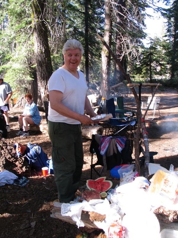 Chuck cooking at the Kegger campsite — smiling while handling foil-wrapped food over a camp fire
