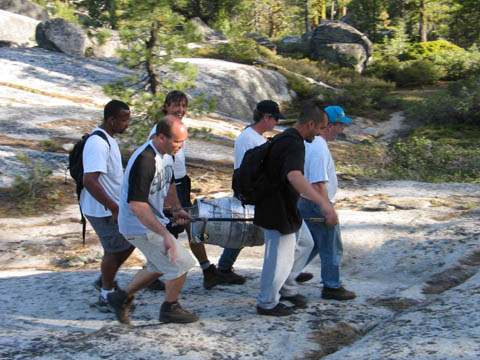 Six men carrying a keg on a steel stretcher across granite slabs